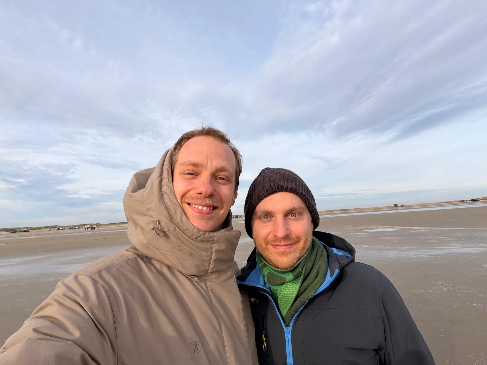Tobi und Jan stehen vor einem breiten Strand und lächeln freundlich zum Betrachtenden 